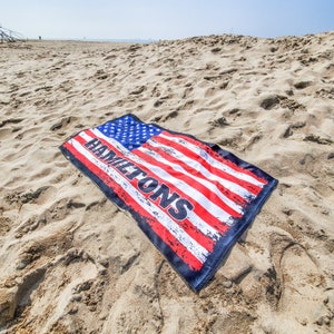 May include: A beach towel with a distressed American flag design and the word "Hamiltons" printed in black across the flag. The towel is lying on a sandy beach.