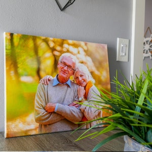 Op de afbeelding: Een canvas print met een portret van een ouder koppel in een parksetting. De man draagt een lichtbruine trui en een bril, en de vrouw draagt een bruine trui en een sjaal. Ze lachen en kijken naar de camera.