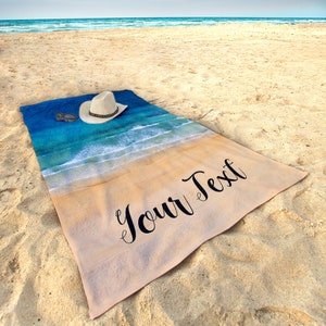 May include: A beach towel with a blue and white ocean scene and a tan sand bottom. The towel has the text "Your Text" printed on the bottom half. A white cowboy hat is lying on the towel.