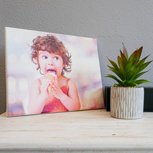 Op de afbeelding: Een canvas print met een jong kind met krullend bruin haar dat een ijsje eet. Het kind draagt een rood-witte jurk en kijkt naar de camera. De achtergrond is wazig en onscherp.