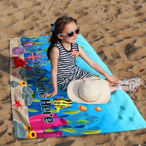 May include: A blue beach towel with a colorful underwater scene featuring fish, coral, and a crab. The text "Your Name Here" is printed on the towel. A young girl is sitting on the towel wearing sunglasses and a striped dress.