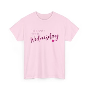 Può includere: Una t-shirt a maniche corte rosa chiaro con il testo rosa scuro "This is what I wear on Wednesday" scritto in una combinazione di caratteri semplici e corsivi, accompagnato da una piccola icona a forma di cuore rosa scuro sotto la parola Wednesday.