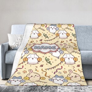 Peut inclure: Un plaid crème doux orné d'un motif répétitif de lapins de dessin animé tenant des pompons, des serpentins colorés et des fanions. Une étiquette centrale en forme de nuage avec une bordure rose contient le mot NAME en lettres bleues.