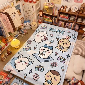 May include: A light blue fleece blanket featuring a repeating pattern of cartoon characters, cameras, ice cream cones, and hearts. The top section includes a camera icon and the text NAME in bold blue letters. The design is printed on a soft fabric.