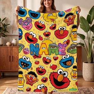 May include: A yellow fleece blanket featuring a repeating pattern of Sesame Street characters including Elmo, Cookie Monster, Big Bird, Bert, Ernie, Abby Cadabby, and Oscar the Grouch. The center displays the word NAME in colorful, bubble-style letters.