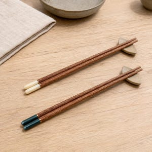 Peut inclure: Deux paires de baguettes en bois reposant sur de petits supports en céramique gris clair incurvés. Une paire présente des extrémités couleur crème, tandis que l'autre a des extrémités bleu sarcelle foncé. Chaque paire a un fin liseré doré.