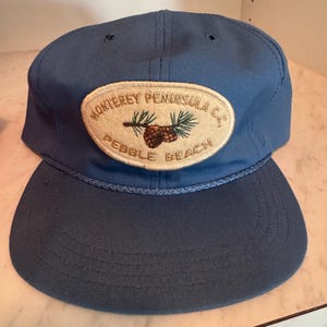 May include: A blue baseball cap featuring a cream-colored oval patch on the front. The patch displays an embroidered image of pine cones and needles with the text "MONTEREY PENINSULA C.C." and "PEBBLE BEACH" in brown lettering. A blue cord trim.
