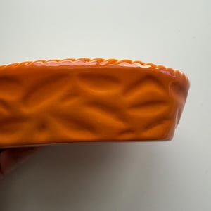 May include: A bright orange ceramic pie dish with a glossy finish. The exterior features a textured, molded pattern resembling a woven basket weave design, and the top rim has a scalloped, crimped edge detail consistent with traditional pie crusts.