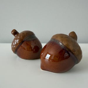 May include: Two ceramic acorn-shaped shakers with a glossy brown finish. Each piece features a textured cap in a lighter tan shade and a smooth, rounded base in a darker brown glaze, with a small stem protruding from the top of each acorn.