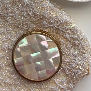 Può includere: Uno specchietto da borsa rotondo con intarsio in madreperla a scacchi e cornice in metallo dorato, appoggiato su una borsa color crema ricoperta di perline bianche e paillettes, con una piccola parte di un piattino a motivi floreali visibile.