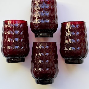 May include: A set of four deep red glass tumblers, each featuring a distinct raised circular bubble pattern across the exterior surface. The glasses have a cylindrical shape with a slightly rounded base and a smooth, open top rim for drinking.