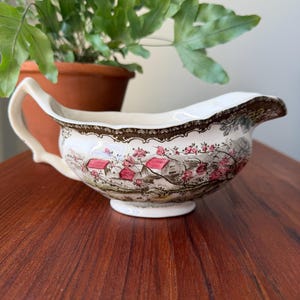 May include: A cream-colored ceramic gravy boat featuring a detailed transferware landscape scene with small red-roofed houses, green foliage, and pink floral accents. The vessel has a curved handle, a wide pouring spout, and a decorative brown border.