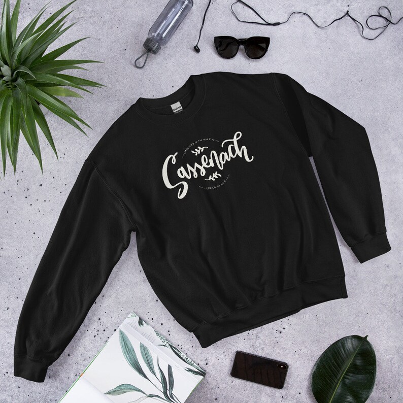 Black crew neck sweatshirt with hand lettered design that reads Sassenach , established in the year 1743, Craigh na Dun and shows two laurels above and below the lettering.