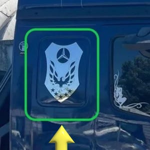 Può includere: Un adesivo color argento a forma di scudo applicato sul finestrino scuro di un veicolo. L'adesivo presenta un logo Mercedes-Benz centrale in alto, un'aquila stilizzata al centro e una fila di cinque stelle lungo il bordo inferiore dello scudo.