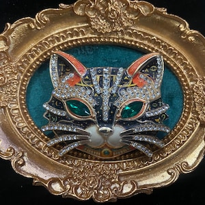 May include: A cat-shaped brooch featuring a dark blue enamel base with gold-tone accents, clear rhinestones, and two large green marquise-cut stones for eyes. The brooch is displayed inside an ornate, oval-shaped gold-tone metal frame.