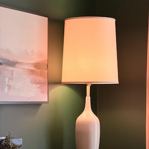 May include: A table lamp with a smooth, off-white ceramic base and a cylindrical, light-colored fabric lampshade. The lamp emits a warm, soft glow, illuminating the area around it. The base features a tapered neck and a metallic circular bottom.