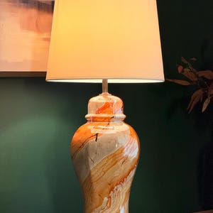 May include: A table lamp featuring a ceramic base with a glossy, marbled finish in shades of orange, cream, and brown. The lamp has a cylindrical, off-white fabric lampshade and a circular metallic base in a brushed gold finish.