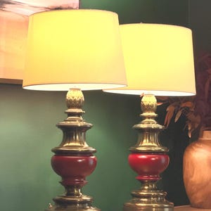 May include: A pair of table lamps featuring brass bases with a central red painted section, topped with off-white drum shades. The lamps have a tiered, ornate metal structure and a small switch on the base, emitting a warm, soft light.