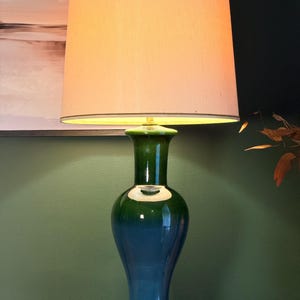 May include: A table lamp with a tall, tapered off-white fabric shade and a glossy ceramic base. The base features a smooth, gradient finish that transitions from a deep forest green at the neck to a vibrant teal blue at the bottom, set on a gold base.