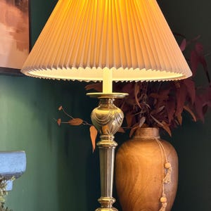 May include: A table lamp with a polished brass base featuring a tiered, fluted column design and a wide, circular foot. The lamp is topped with a cream-colored, pleated fabric lampshade that emits a warm, soft glow when illuminated from within.