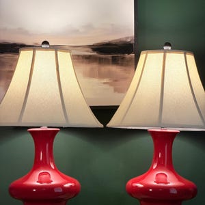 May include: A pair of table lamps featuring glossy red ceramic bases with a wide, rounded bottom and a narrow neck. Each lamp has a square black base and a flared, off-white fabric lampshade with a clear finial at the top of the shade structure.