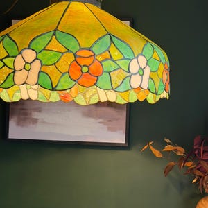 May include: A stained glass pendant lamp featuring a vibrant yellow background with a floral pattern of orange and white flowers surrounded by green leaves. The lamp has a scalloped edge and a dark metal frame holding the glass pieces together.