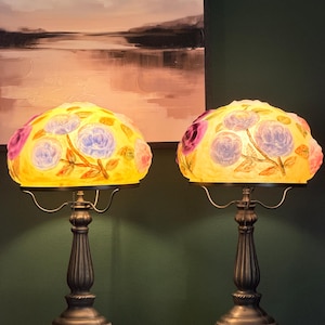 May include: A pair of table lamps featuring dome-shaped glass shades with a yellow background and painted purple and blue floral patterns. The lamps have dark metallic, fluted pedestals with ornate, wide bases and pull-chain switches.