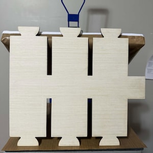 May include: A light-colored, flat wooden cutout featuring a rectangular central body with three protruding tabs along the top edge and three matching tabs along the bottom edge, plus a single rectangular extension on the right side.