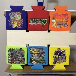 May include: Six foam can coolers in purple, red, orange, lime green, light blue, and yellow. The purple cooler shows crawfish boil graphics. The red one reads Grand Daddy's Smokehouse Barbecue. The orange one says Blame it on the Drink Package. The lime green one features Louisiana imagery. The light blue one depicts a Louisiana map. The yellow one displays an LSH Tigers mascot.