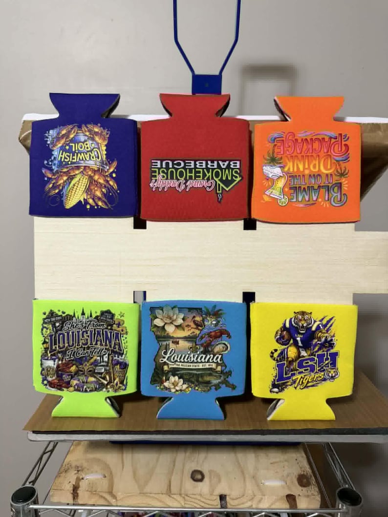May include: Six foam can coolers in purple, red, orange, lime green, light blue, and yellow. The purple cooler shows crawfish boil graphics. The red one reads Grand Daddy's Smokehouse Barbecue. The orange one says Blame it on the Drink Package. The lime green one features Louisiana imagery. The light blue one depicts a Louisiana map. The yellow one displays an LSH Tigers mascot.