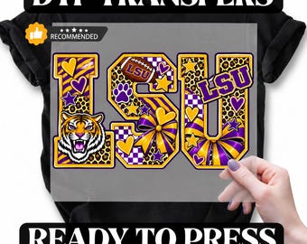 May include: A DTF transfer sheet featuring the letters LSU in purple and gold, decorated with tiger stripes, leopard print, checkered patterns, hearts, stars, a football, a tiger head, and bows, positioned on a black t-shirt for heat pressing.