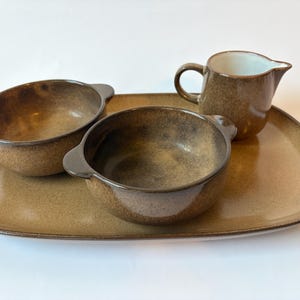 May include: A ceramic set consisting of a large rectangular tray with rounded corners, two small round bowls with side handles, and a small pitcher with a handle and spout. All pieces feature a speckled brown glaze with darker edges.