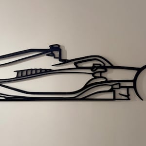 May include: A black, flat, wall-mounted silhouette of a Formula One race car. The design features the distinct aerodynamic profile of an open-wheel vehicle, including the front wing, cockpit area, side pods, and a large rear wing assembly.