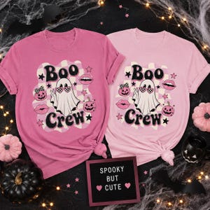 May include: Two short-sleeved t-shirts in shades of pink featuring a graphic print with the text "Boo Crew". The design includes a ghost wearing heart-shaped glasses, pumpkins, lips, and stars set against a checkered background pattern.
