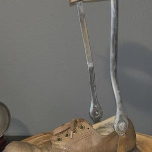 May include: A medical leg brace featuring a tan leather cuff with a metal buckle and two vertical silver metal support bars. The bars are attached to a tan leather oxford shoe with brown laces, featuring a low heel and a rounded toe design.