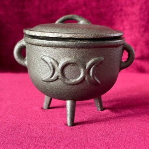 May include: A small, dark gray cast iron cauldron with a matching lid, three sturdy legs, and two side handles. The front of the cauldron features a raised triple moon symbol, consisting of a central circle flanked by two crescent moons.
