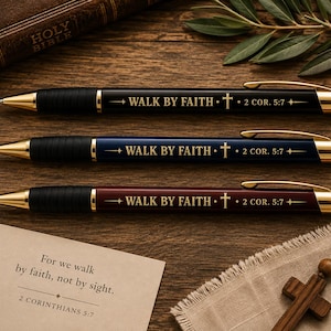 Könnte beinhalten: Drei einziehbare Kugelschreiber in Schwarz, Marineblau und Kastanienbraun, jeweils mit goldfarbenen Akzenten, einem schwarzen Griff und dem eingravierten Text WALK BY FAITH mit einem Kreuzsymbol und der Referenz 2 COR. 5:7 auf dem Schaft.