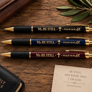 May include: Three retractable ballpoint pens in black, navy blue, and maroon, each featuring gold-tone clips, tips, and accents. The barrels are engraved with the text BE STILL, a cross symbol, and PSALM 46:10, flanked by leaf motifs.