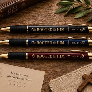 Könnte beinhalten: Drei einziehbare Kugelschreiber in Schwarz, Blau und Burgunderrot, jeweils mit goldfarbenen Akzenten, einem schwarzen Griff und dem aufgedruckten Text ROOTED IN HIM mit einem Kreuzsymbol und der Referenz COL. 2:7, begleitet von einer Karte.