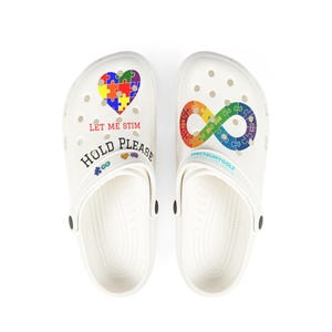 May include: A pair of white clogs featuring a colorful puzzle piece heart and the text LET ME STIM HOLD PLEASE on the left shoe, and a rainbow infinity symbol made of puzzle pieces on the right shoe, with small icons and the text SPECTRUMTOOLZ.