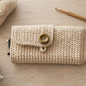 Puede incluir: Un estuche rectangular de ganchillo en color beige con un patrón de puntos texturizados, un cierre de solapa asegurado por un botón de madera marrón oscuro y una pequeña etiqueta de tela blanca en el lateral con la palabra HANDMADE.