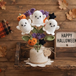 May include: A crochet bouquet featuring white ghosts, orange pumpkins, purple bats, and black stars on green stems in a speckled ceramic vase. A round tag attached to the vase reads Spooky Blooms Crochet. The arrangement sits on a white doily.
