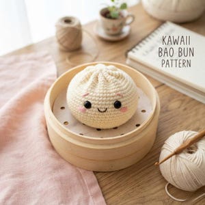 May include: A cream-colored crocheted bao bun with black bead eyes, a stitched smile, and pink embroidered cheeks. The plush bun sits inside a round bamboo steamer basket. Nearby are a ball of yarn, a crochet hook, and a notebook with the text KAWAII BAO BUN PATTERN.