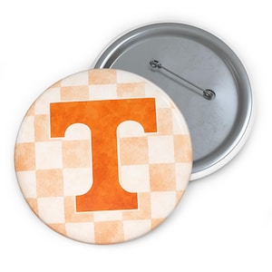 May include: A round pin-back button featuring a watercolor-style orange letter T centered on a light orange and white checkered background. The back of the button is silver-toned metal with a horizontal pin mechanism for secure attachment.