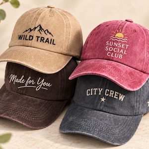 May include: Four baseball caps with a distressed finish. The tan cap features a black mountain graphic and the text WILD TRAIL. The maroon cap displays a yellow sun and waves with SUNSET SOCIAL CLUB. The dark brown cap says Made for You. The dark gray cap features CITY CREW and two stars.