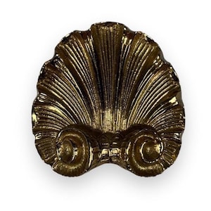 May include: A metallic gold decorative object shaped like a scallop shell with prominent vertical ridges radiating from the center. The base features two symmetrical, tightly coiled spiral elements that anchor the shell design in a polished finish.