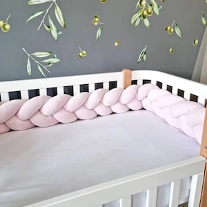 May include: A white crib featuring a soft, light pink braided bumper that lines the interior perimeter. The crib is equipped with a plain white mattress cover, and the surrounding wall displays a pattern of green olive branches and olives.