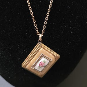 May include: A gold-toned metal locket pendant shaped like a small book, suspended from a matching gold-toned chain. The front of the locket features a rectangular white inlay decorated with a small pink rose and green leaves, framed by a textured border.