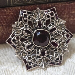 May include: A silver-toned brooch featuring a star-shaped filigree design with deep red enamel accents and small, clear embedded crystals. A large, square-cut, dark red gemstone is set in the centre of the piece, surrounded by intricate metalwork.