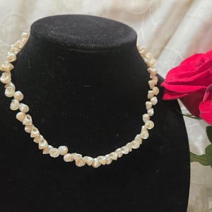 Puede incluir: Un collar compuesto por numerosas perlas blancas pequeñas e irregulares ensartadas en un hilo circular continuo. Las perlas presentan una superficie lustrosa y texturizada con formas orgánicas variadas, creando un aspecto denso y superpuesto.